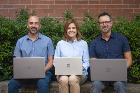 Lee Chandler, Krista Bulger, and Joe Dlugo sit smiling on a bench with open laptops
