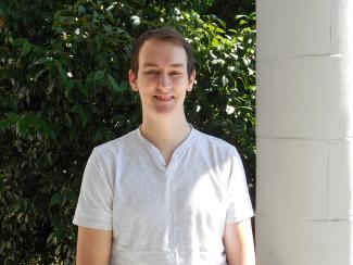 Joel is smiling and standing outside of the old main building on a sunny day