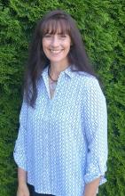 Head shot of Carmina Harrington against a row of green arborvitaes. A middle-aged woman with long brown hair smiling happily.