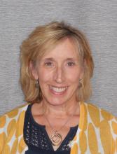 Gretchen Gianotti, she has light medium length hair and a happy smile. She is wearing a sunny yellow sweater and blue dress.