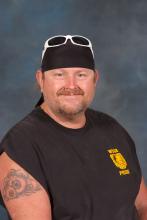 John Fleming, a smiling, blue eyed, bearded and mustached man, wearing a black WSSB t-shirt, black biker cap with sunglasses.