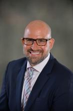 Middle-aged, white male with a bald head, short beard, and glasses wearing a dark blue suit and proudly smiling 