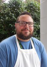 William Carroll short  dark brown hair short dark beard wearing a cooks apron and a blue shirt. With a full smile.