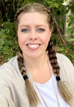 Jackie Tucker, a woman with brown/blonde hair in two braids, happily smiling wearing a white shirt and tan sweater.