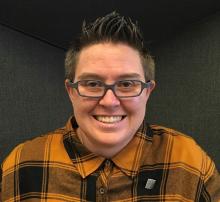 Jesse Bolt is smiling in a gold/black plaid button down shirt, wearing glasses and a they/them pronoun pin on their shirt.