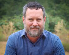 A man in his early 40’s with brown eyes and graying hair and beard amidst a garden backdrop.