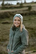 Tracy Spohn smiling while standing on grassy slope in the Columbia River Gorge; pine trees and river in background.