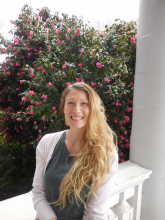 Amanda Pizzo, a smiling woman with big hair. Backdrop is a large, pink flowered tree on a warm summer day.