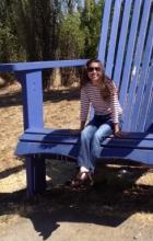 On a giant blue chair, Khanh is a woman with dark hair and sunglasses sits smiling.
