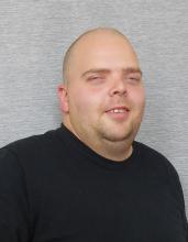 Matt Hines, a mid-30’s male, with short hair, in a black t-shirt, sitting with some swag and smiling big.