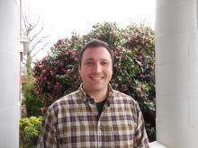 Head shot of Sam Schrager smiling. Brown hair and eyes, plaid shirt.