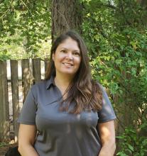 Melinda Bakos, a smiling woman in her 40’s wearing a gray shirt and standing in front of a tree.