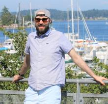 Paul wears sunglasses and a hat while posing in front of a sailboat filled marina.