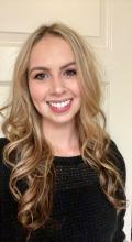 Lisa “Elle” Horton, a woman in her mid 20’s with hazel eyes and long blonde hair that is curled, smiling happily wearing a black knit sweater.
