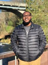 Josh, a smiling man with his hands in the pockets of his jacket standing on a bridge in front of Tumwater Falls.