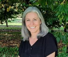 Lindsey is smiling at the camera, wearing a black top, has long gray hair, and silver hoop earrings. The background is a cherry orchard, the trees full of green leaves. 