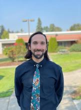 Sean, a middle aged white male with shoulder length hair and beard wearing a navy shirt and floral tie standing outdoors smiling on a sunny day.