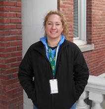 Anita is smiling with her hands in her jacket pockets standing outside of the old main building.