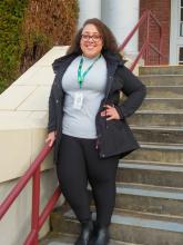 Kayla is smiling while standing posed on the stairs with her hand on her hip while holding the the stair railing. She has really curly brown hair and wears glasses