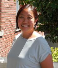 A medium skin-toned Asian woman smiles at the camera. She wears earrings, a light grey blouse, and has chin length hair that is tucked behind one ear.