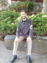 Bearded, smiling, and wearing khakis and a dark grey button-up—sits on a bench in the sun in front of a bush in his first week as the new Procurement Specialist at WSSB.